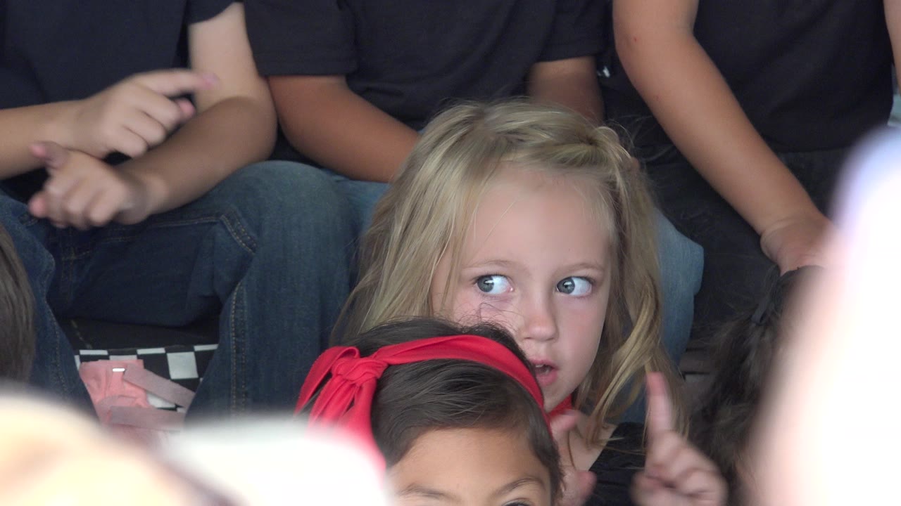 Blonde child seated among other children with a red headband in the foreground.