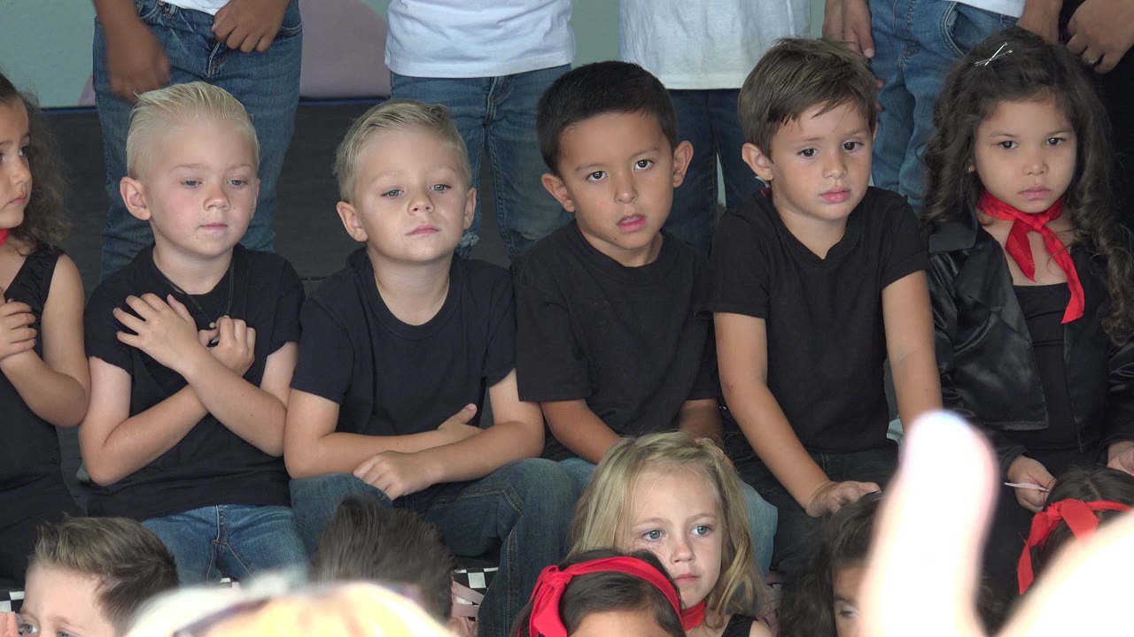 A row of children in Cabo San Lucas, Mexico, remains seated in black shirts while an out-of-focus raised hand and heads partially block the lower foreground.