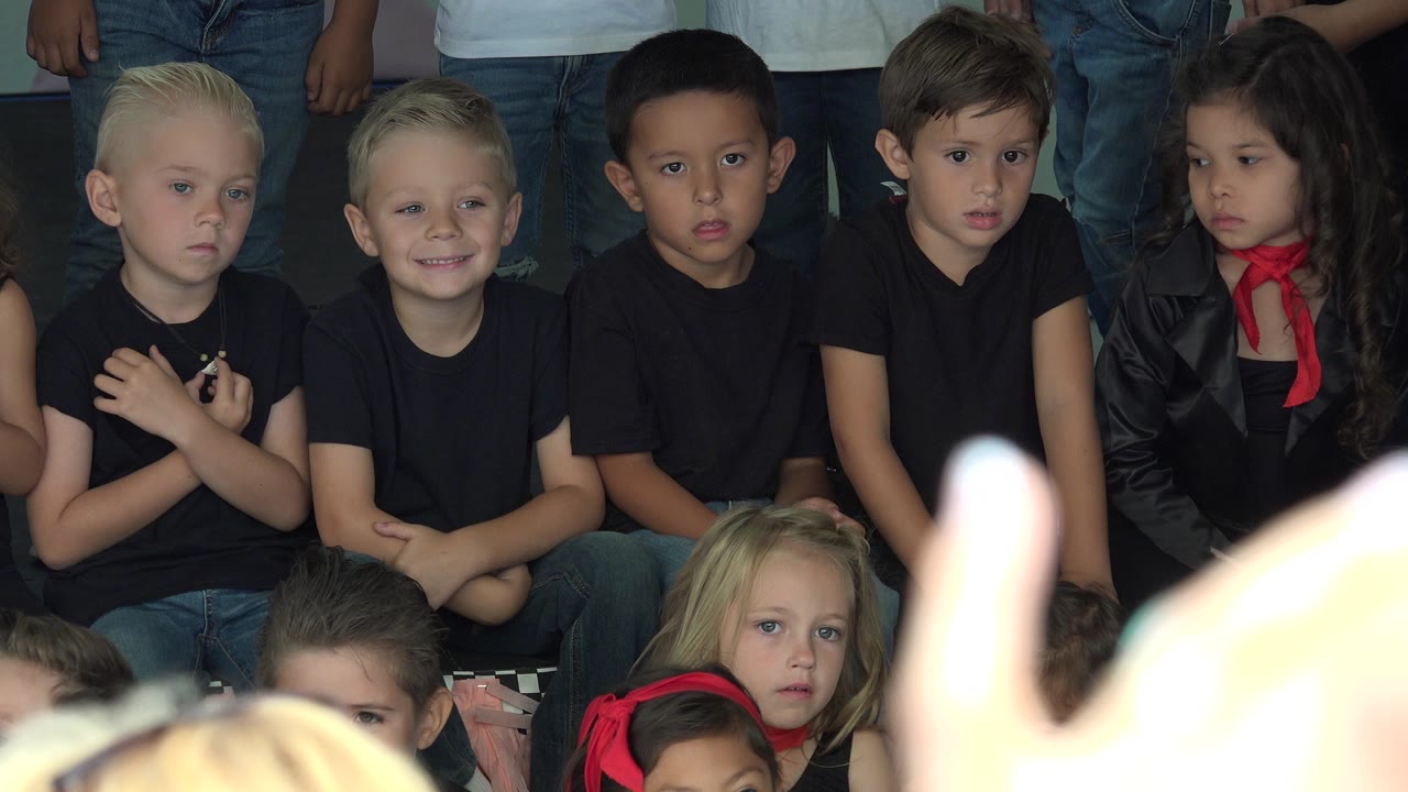 At 0:01, the same group of children remains seated in Cabo San Lucas, Mexico, with one blond boy smiling while others look forward or sideways.