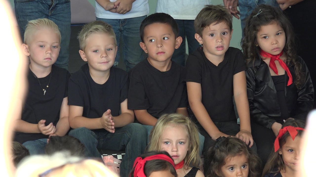 At 0:00 in Cabo San Lucas, Mexico, several young children in black tops sit close together with adults' legs cropped behind them and other children partly visible at the bottom edge.