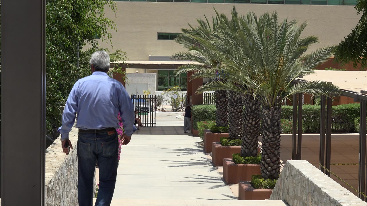 An elderly man in a blue shirt walks away from camera down outdoor steps in Cabo San Lucas, Mexico, with a palm tree and stone walls ahead.