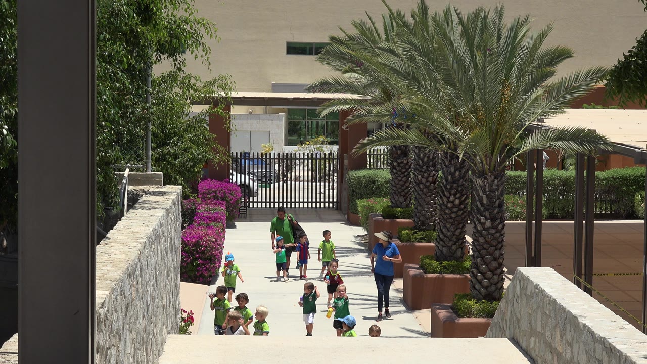 At the start of the Cabo San Lucas clip, children and adults appear on a bright walkway framed by stone walls, pink shrubs, palms, hedges, and a black gate.