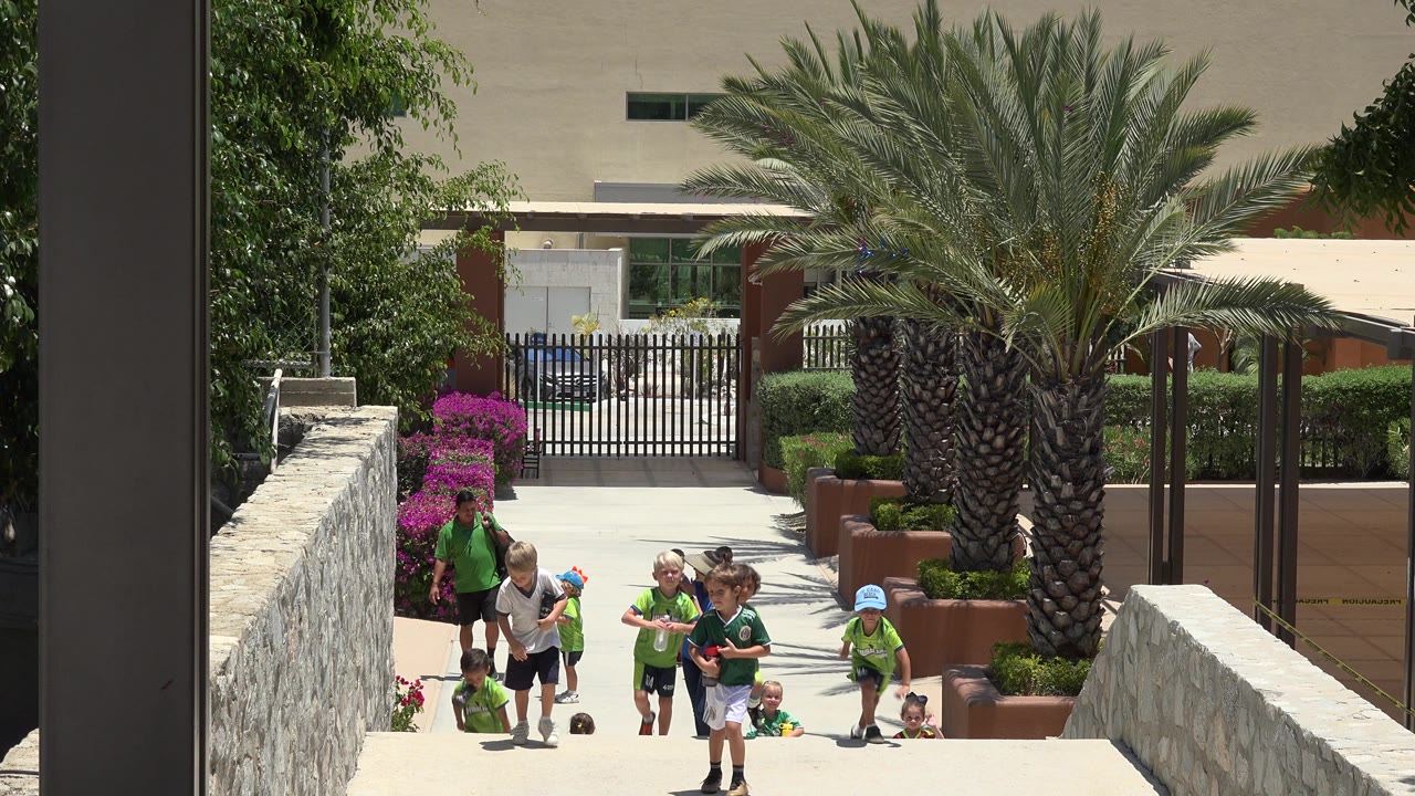 At the start of the Cabo San Lucas clip, children in green shirts and nearby adults move up a bright outdoor walkway lined by stone walls, flowers, and a large palm.