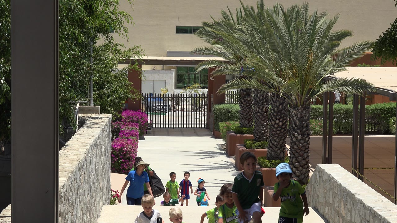 A group of children in soccer uniforms and an adult in a blue shirt move up the walkway in Cabo San Lucas, Mexico, framed by stone walls, landscaping, and a palm tree.