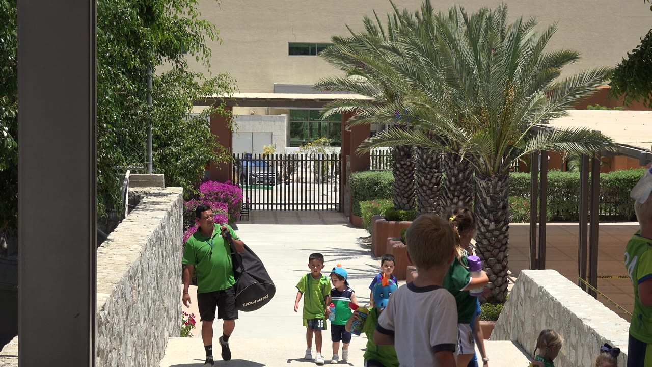 Children and adults on a sunny walkway near a palm tree and building.