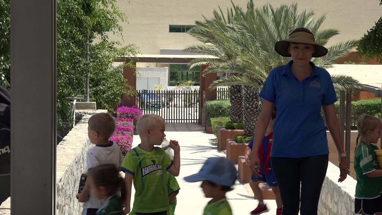 Children in soccer uniforms and a woman in a blue polo on a sunny walkway.