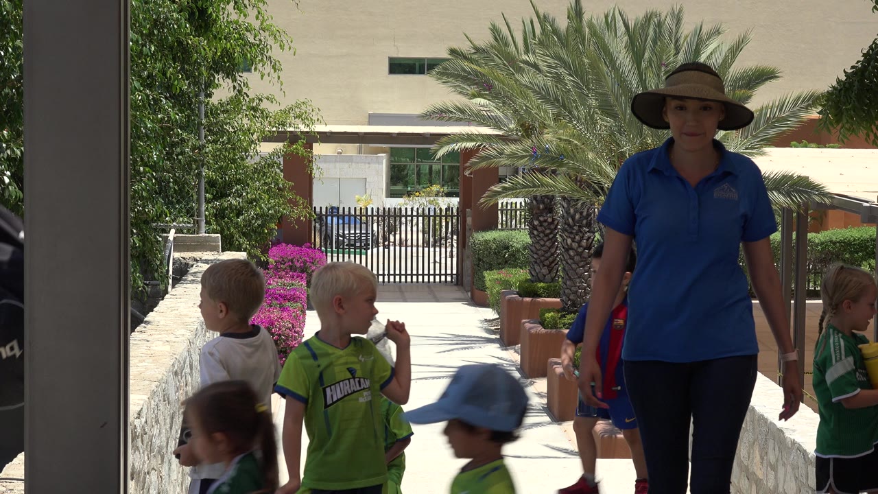 A woman in a blue polo and sun hat remains centered on the shaded edge of the frame while children in soccer uniforms stand and walk behind her in Cabo San Lucas, Mexico.