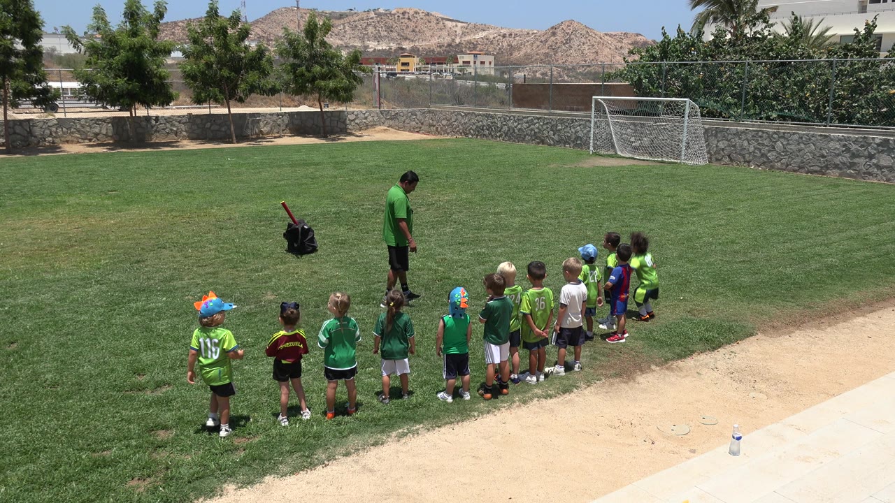 Children lined up on a grass football field with a coach nearby.