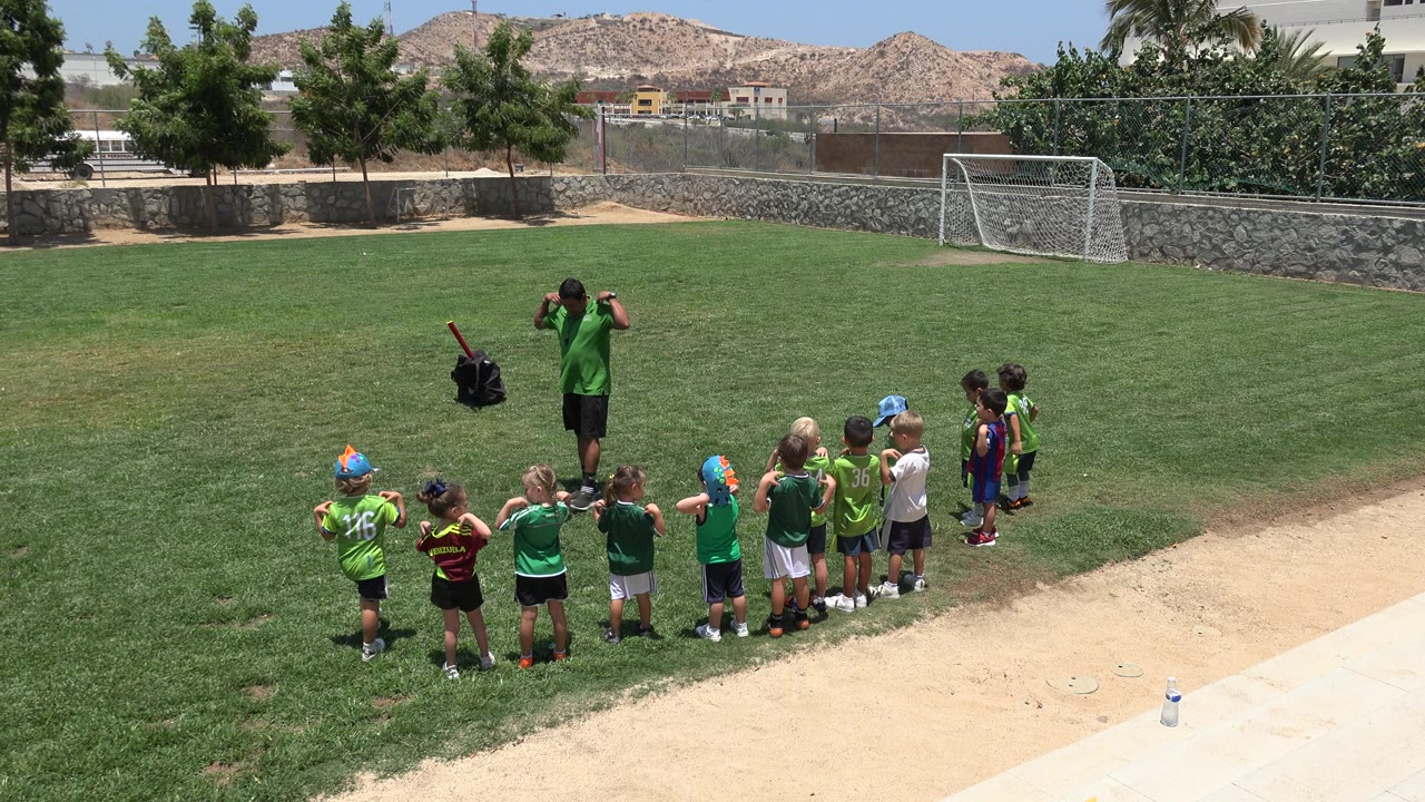 A coach in green faces a row of children doing warm-up motions on a sunny grass field in Cabo San Lucas, Mexico, with a soccer goal, trees, stone wall, and hills behind them.