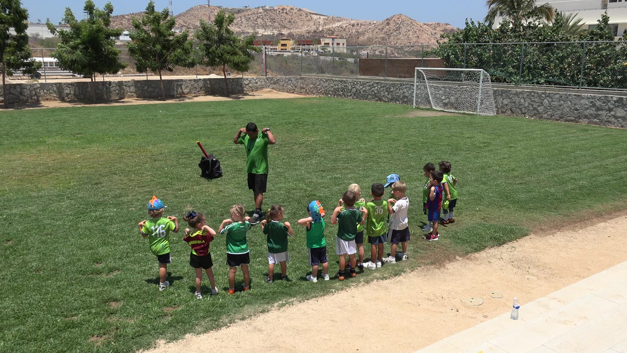 The coach stands near the center of the field with both arms raised as young children in sports outfits face him in a line in Cabo San Lucas, Mexico.
