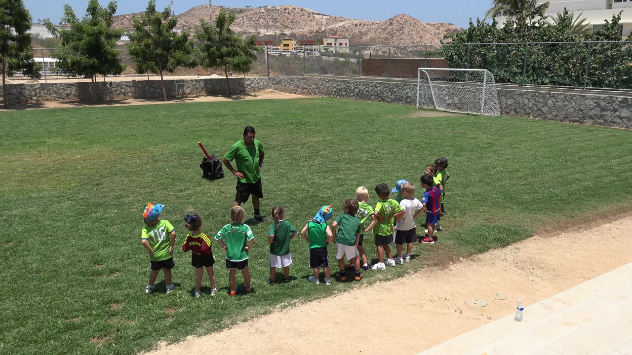 Adult in green shirt stands near children lined up on a grass field.