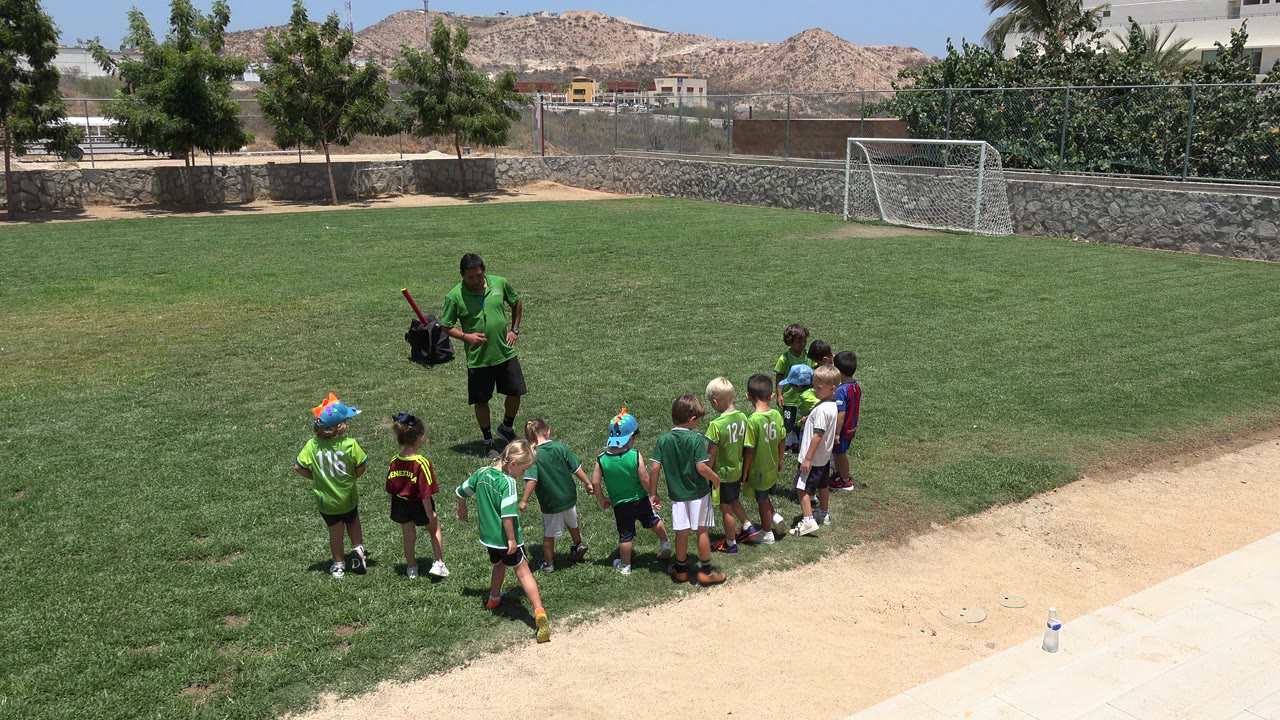 At 0:00 in Cabo San Lucas, a coach in green faces a small group of children on a grass soccer field, with a goal, stone wall, trees, and dry hills behind them.