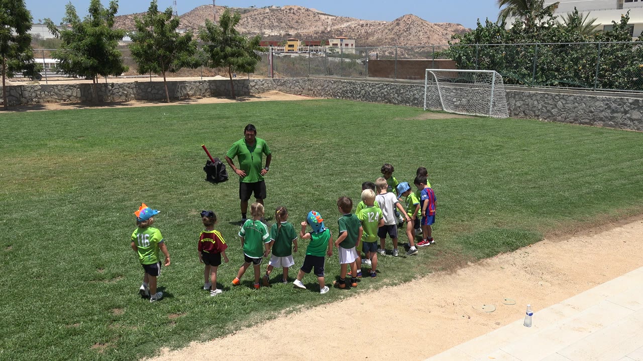 At 0:01, the children remain grouped in front of the green-shirted instructor on the Cabo San Lucas field, with open grass, a white goal, trees, and hillside behind them.
