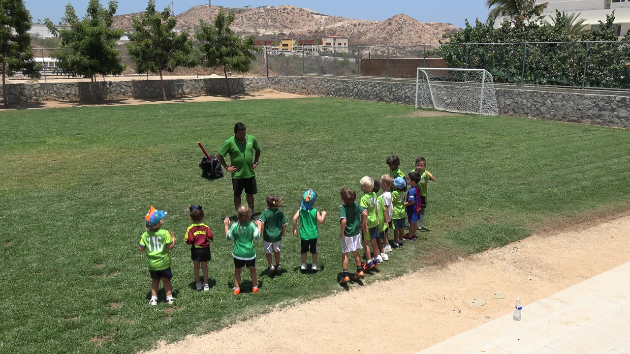 In Cabo San Lucas, a coach in green faces young children lined up on a sunny grass soccer field, with a goal, stone wall, trees, and dry hills behind them.