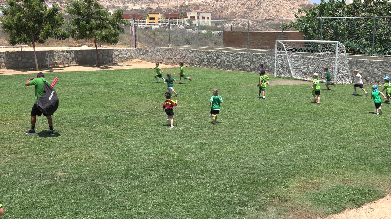 Children in green shirts chase the ball near the goal in Cabo San Lucas, Mexico, while an adult with a large black bag stands in the foreground.
