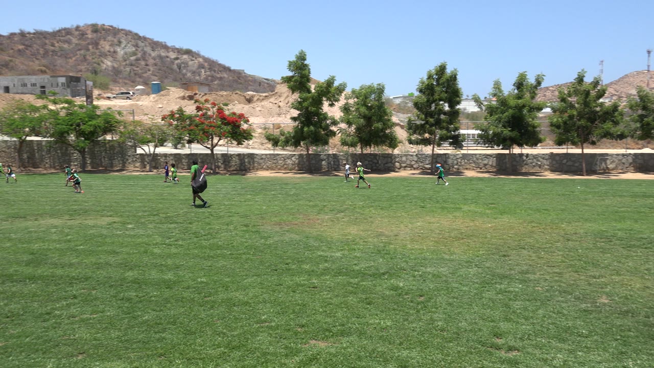 Children in green uniforms move across the grass field in Cabo San Lucas, Mexico, with an adult standing near the play and hillside terrain beyond the wall.