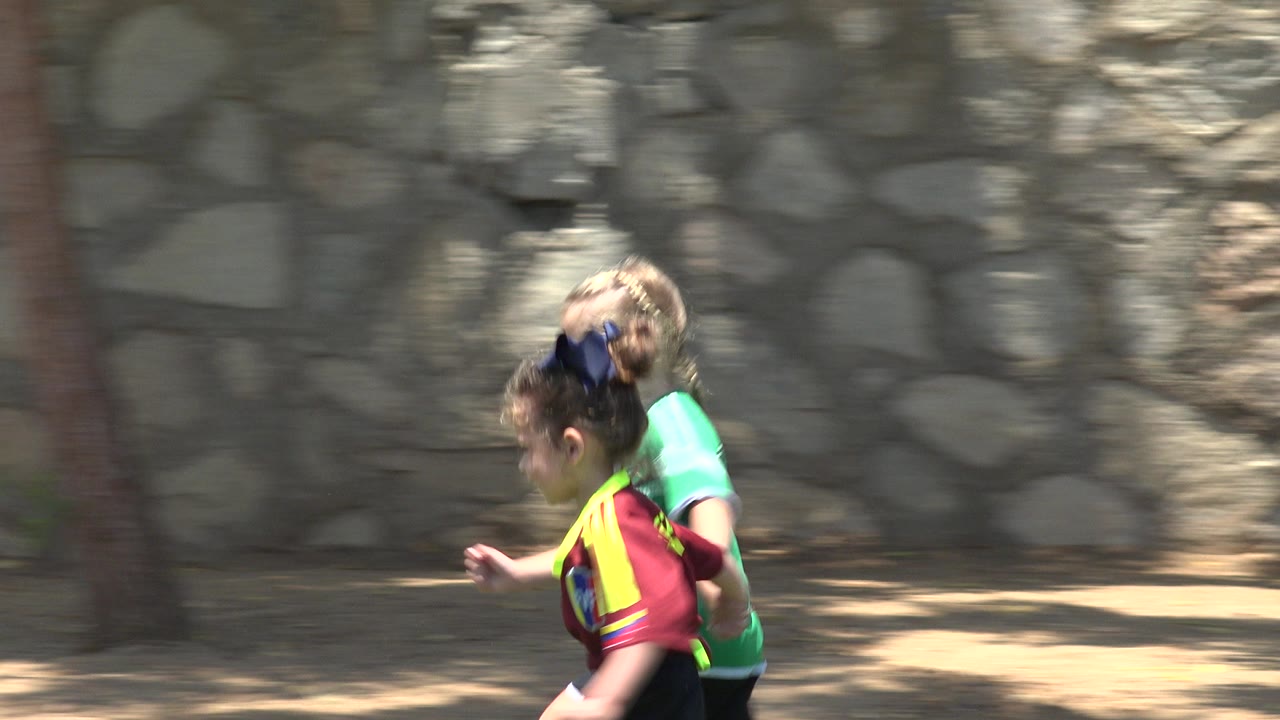 At 0:00, two young girls in bright soccer jerseys run across grass in Cabo San Lucas, Mexico, with a stone wall and shaded dirt behind them.