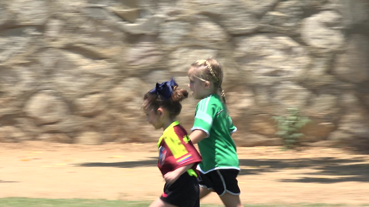 At 0:02, the runners are farther right in the frame in Cabo San Lucas, Mexico, passing a sunlit dirt strip and irregular stone wall.