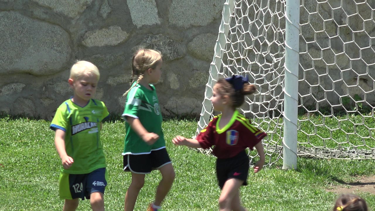 Young children in green and burgundy soccer jerseys stand near a white goal net on a grassy field in Cabo San Lucas, Mexico.