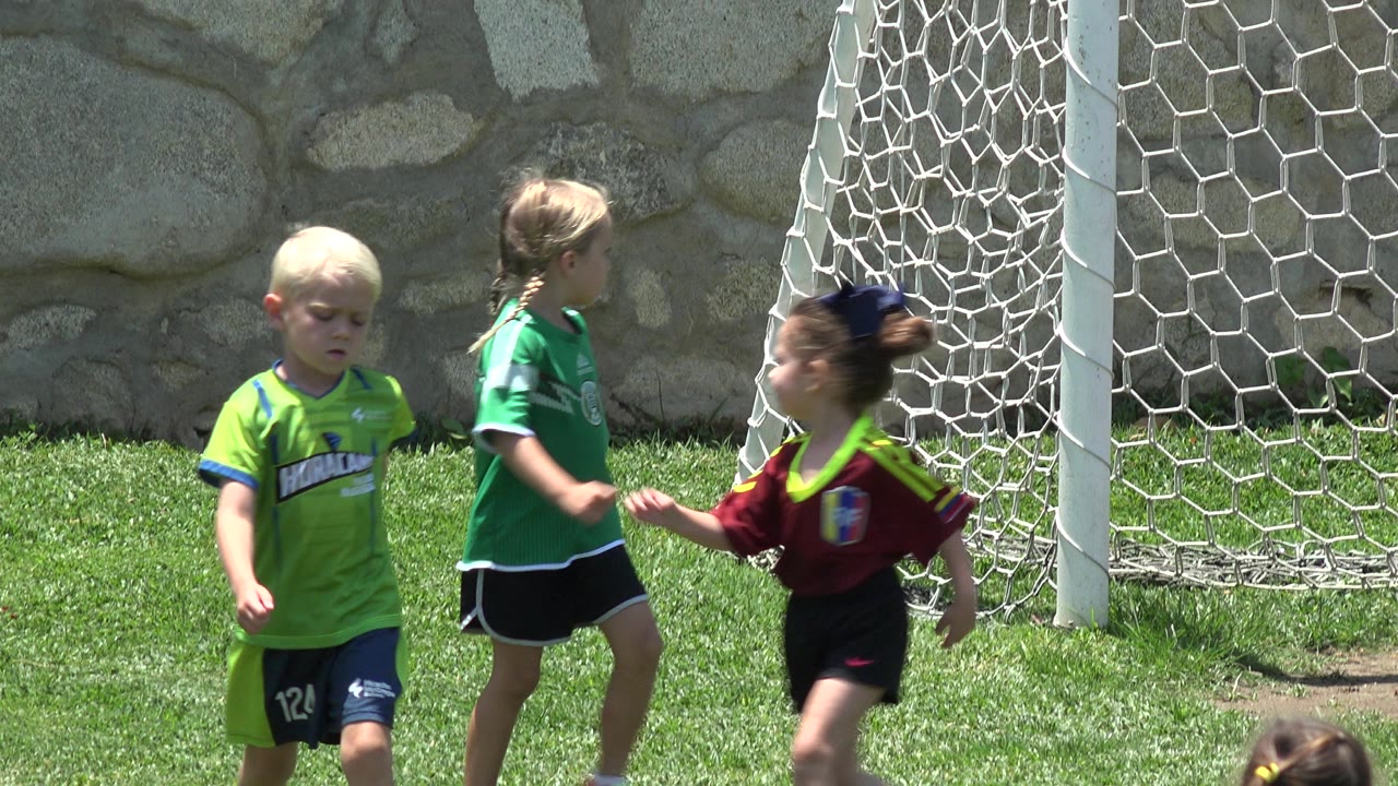Three children pass in front of the goal net, including a blond child in lime green and two children in green and burgundy jerseys in Cabo San Lucas, Mexico.