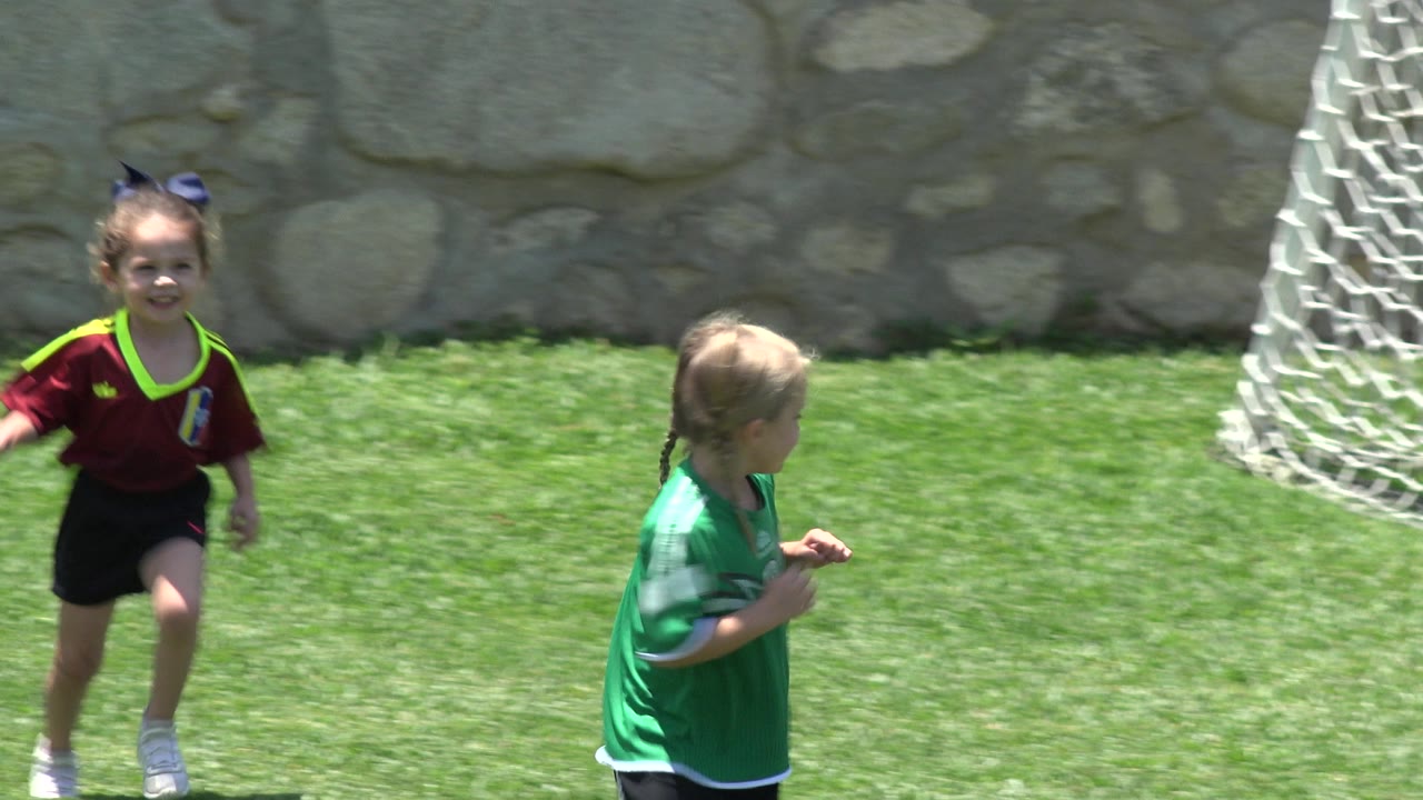 A child in a maroon soccer jersey runs toward camera on a grass field in Cabo San Lucas, Mexico, while a child in green turns away near the goal.