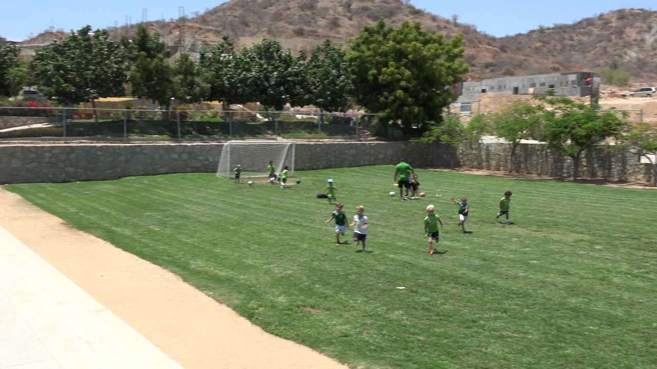 Children play soccer on a green field in Cabo San Lucas, Mexico, with a coach near a small goal, trees, a stone wall, and dry hills in the background.