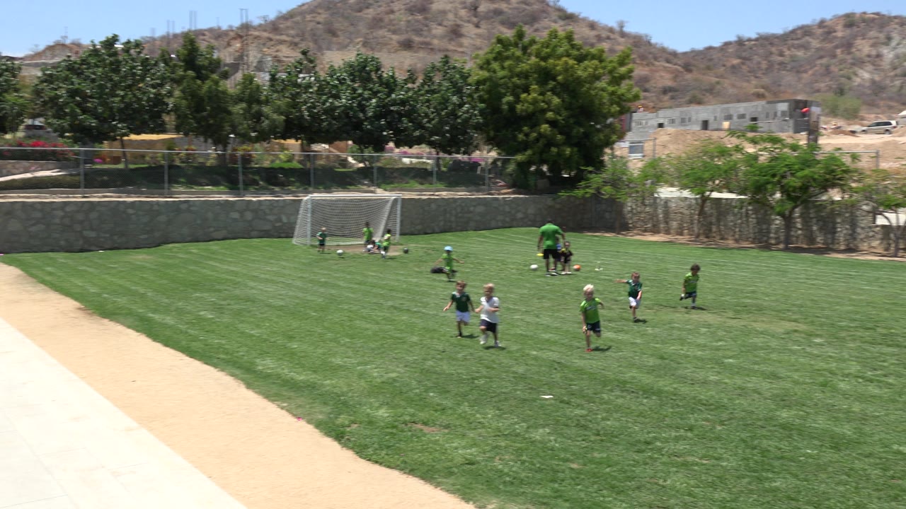 Young children in green jerseys and casual clothes move across the Cabo San Lucas soccer field while the coach remains near balls and a small goal at the far side.