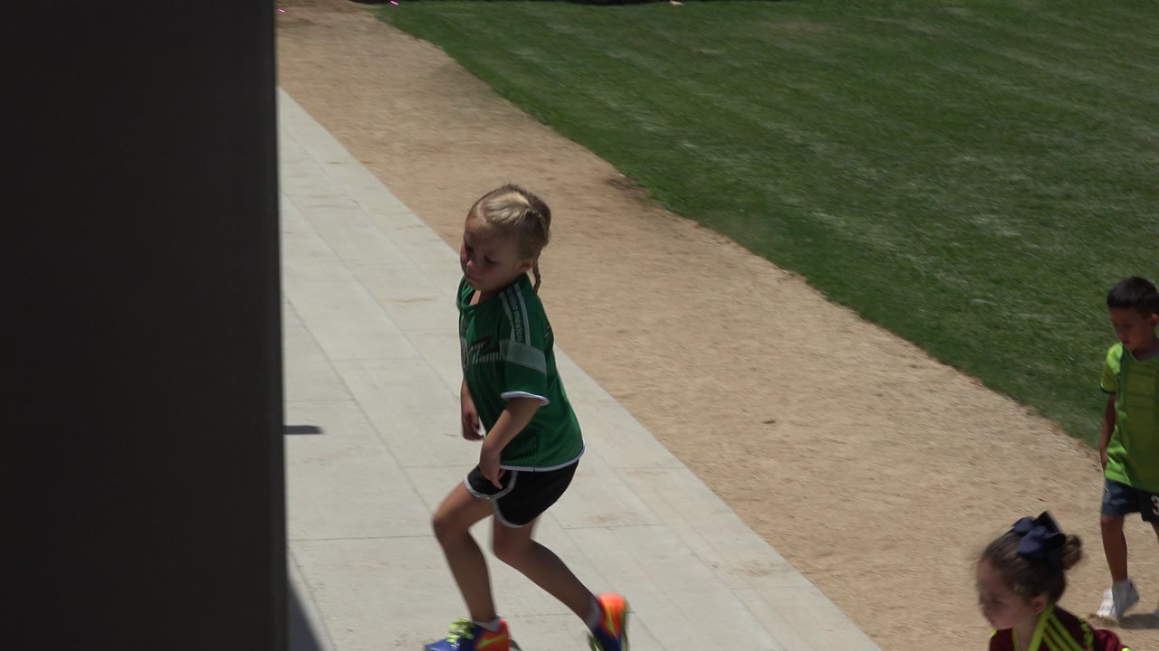 A young player in a green jersey steps along the walkway in Cabo San Lucas while another child stands farther back near the grass and tan dirt strip.