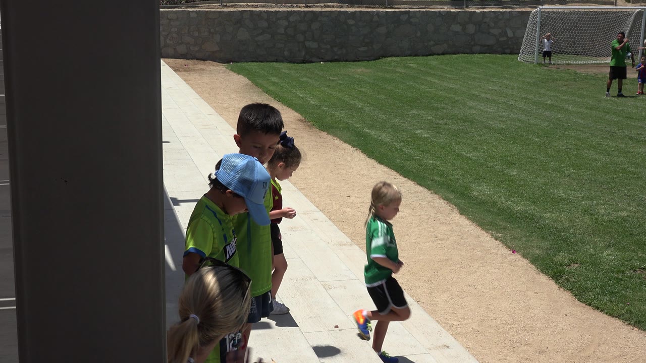 Children in green shirts stand by a walkway beside a grass field.