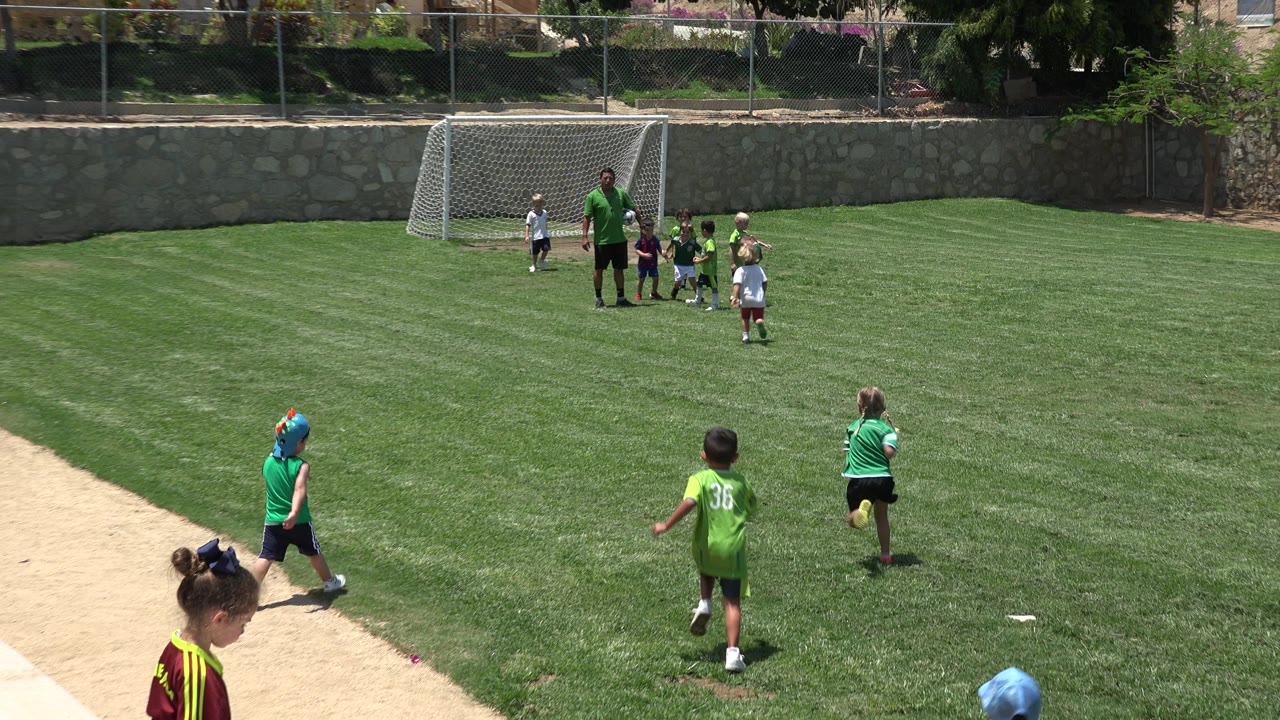 Children in green soccer shirts move across a grass field in Cabo San Lucas, Mexico, while an adult coach stands near a white goal in the distance.