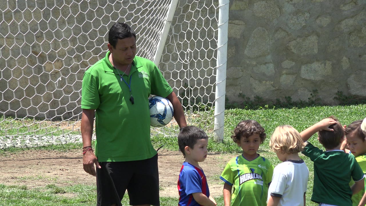 In Cabo San Lucas, Mexico, a coach in a green polo holds a blue and white soccer ball while several children stand near a white goal net in bright sun.