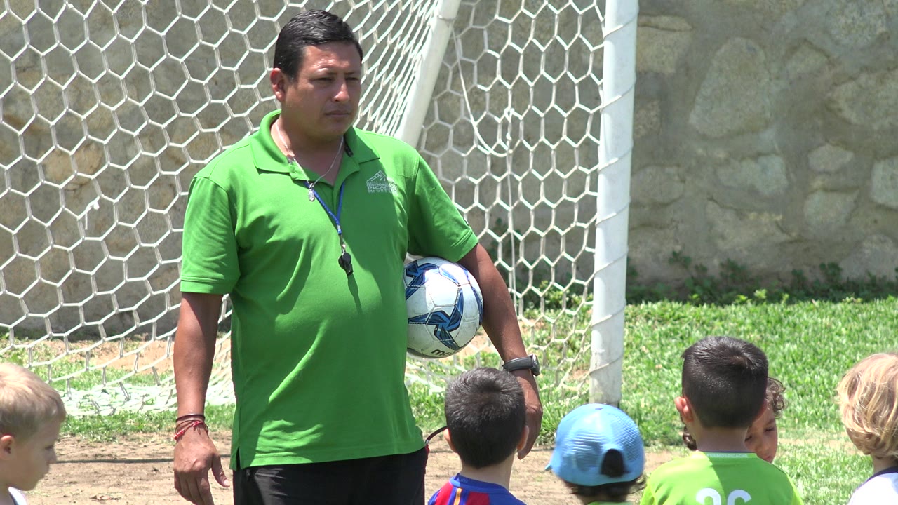 In Cabo San Lucas, Mexico, a coach in a green polo holds a soccer ball near a white goal net while young children stand at the edge of the frame.