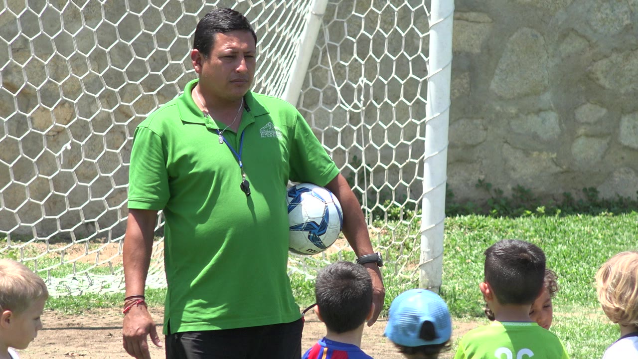 In Cabo San Lucas, Mexico, the coach remains near the goal with the ball tucked at his side while several children in jerseys face him from the foreground.