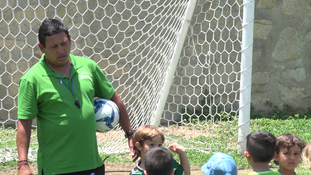 In Cabo San Lucas, Mexico, a coach in a green polo holds a soccer ball beside young children gathered near a white goal net.