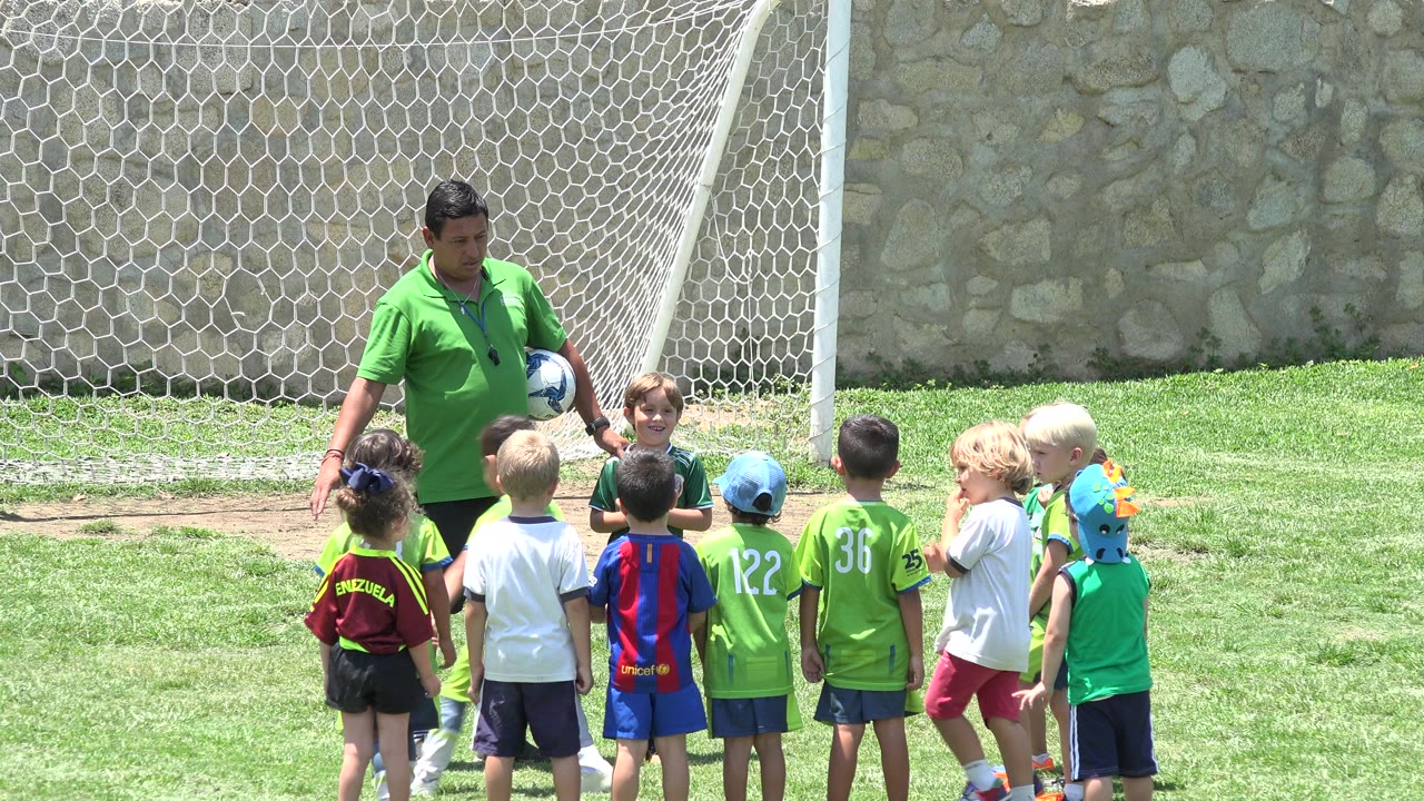 Coach holding a soccer ball in front of children on a grassy field.
