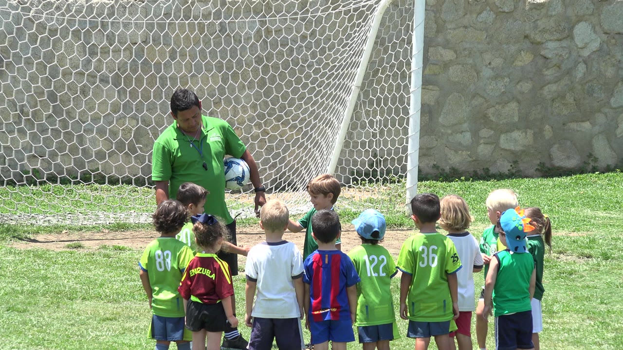 In Cabo San Lucas, Mexico, a coach in a green shirt holds a soccer ball while a group of young children face him near a goal net and stone wall.
