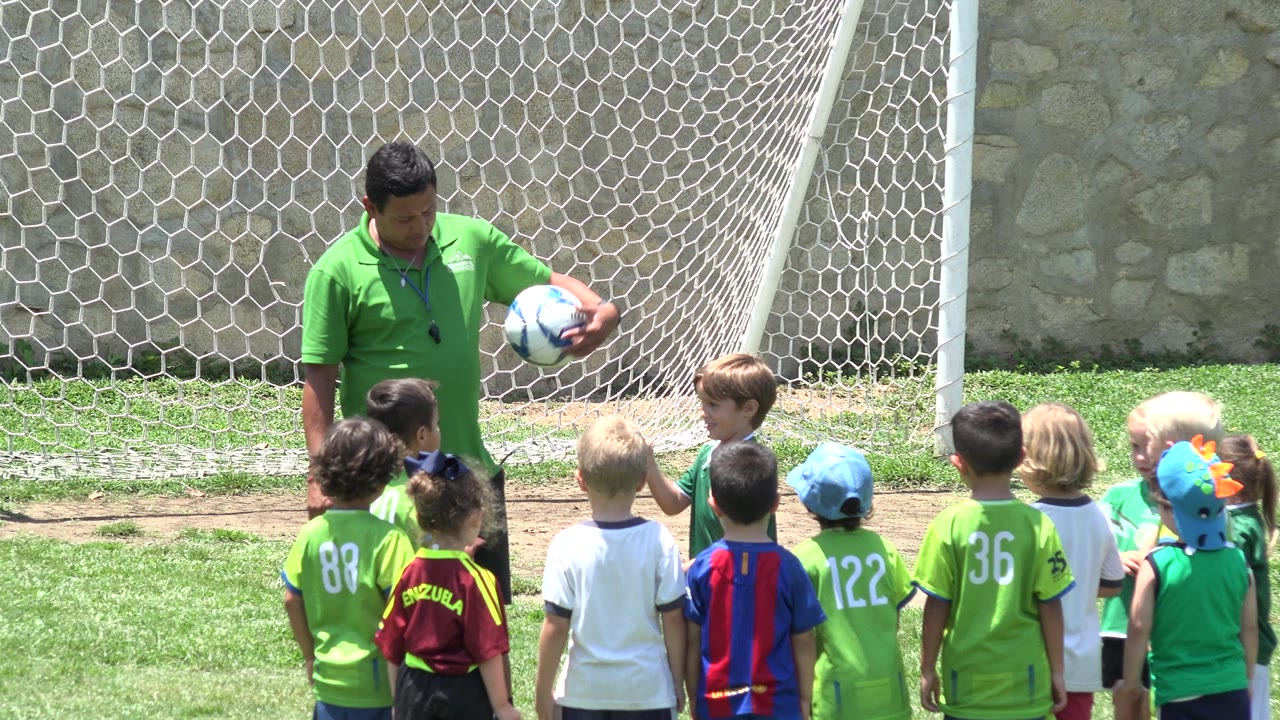Coach gestures with a soccer ball while children face him.