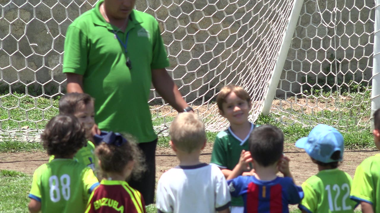 At 0:01 in Cabo San Lucas, a small child in a green jersey looks toward camera as teammates stand with backs turned near the coach and net.