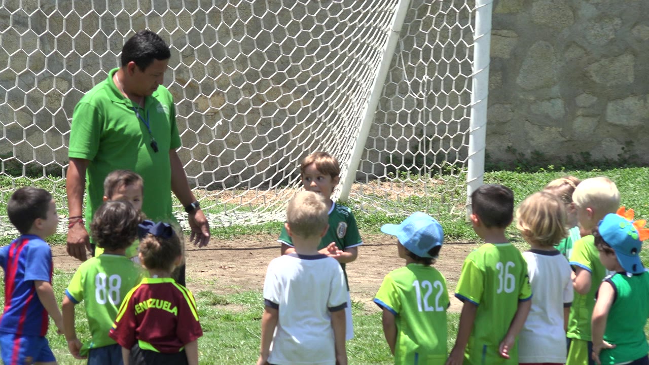 At 0:01 in Cabo San Lucas, Mexico, several children face the coach while a child in a green jersey stands near the goal net and stone wall.