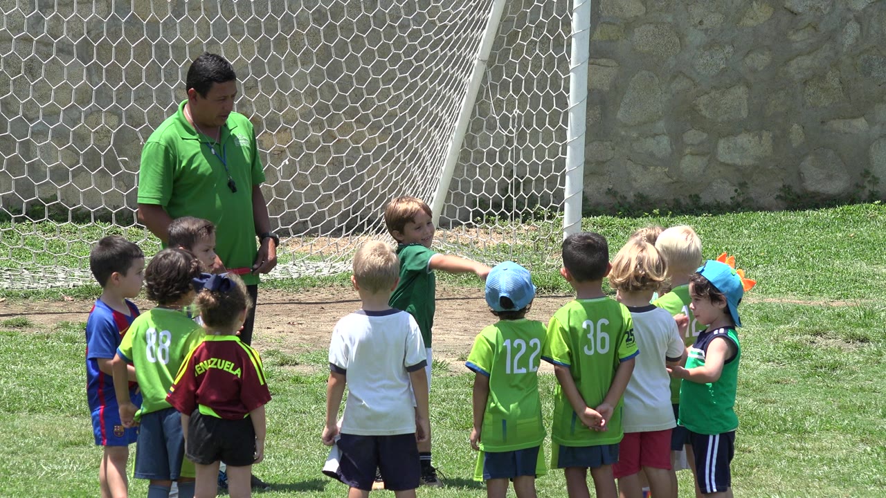 In Cabo San Lucas, Mexico, a child in green points across the clustered group while the coach watches near the soccer goal net.