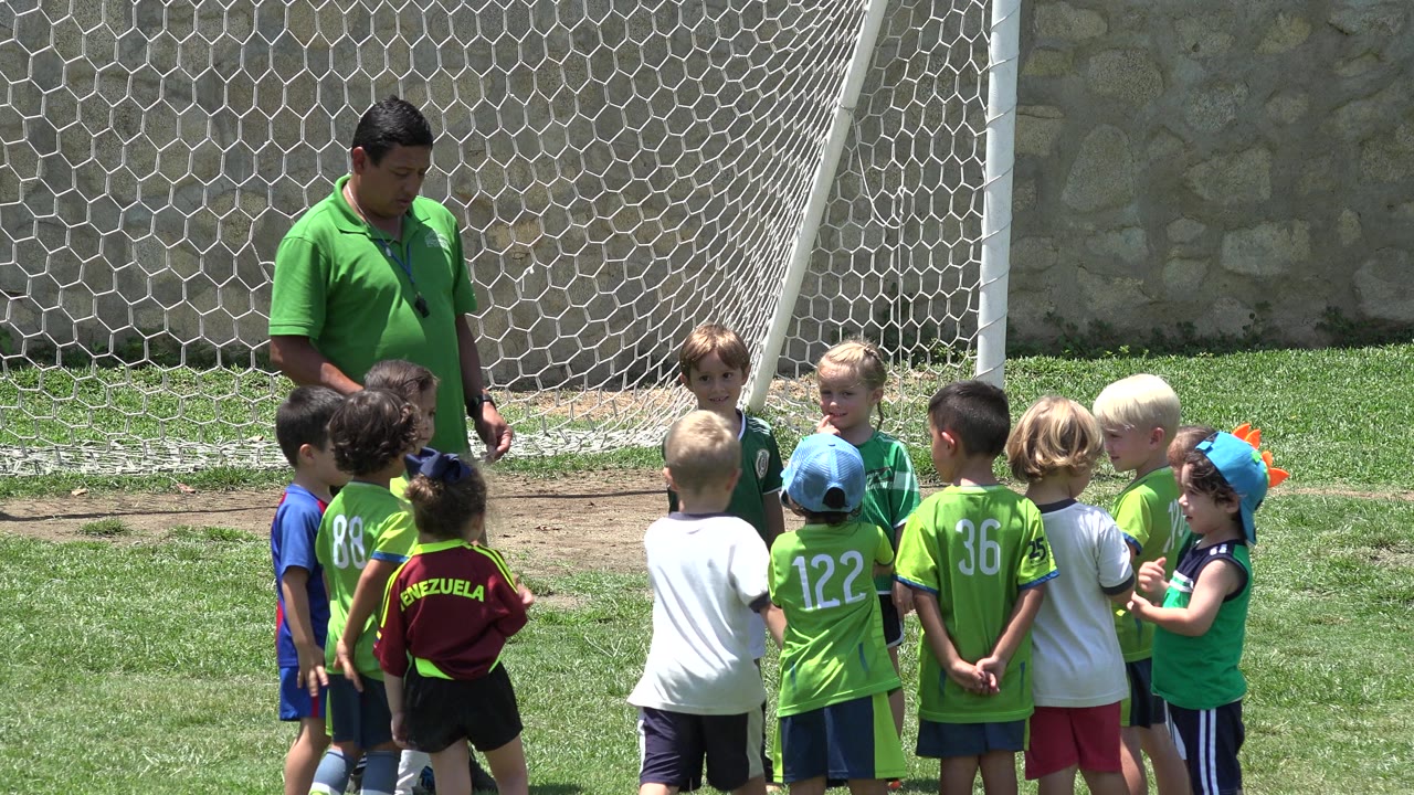 Coach stands beside children gathered near a soccer goal net.