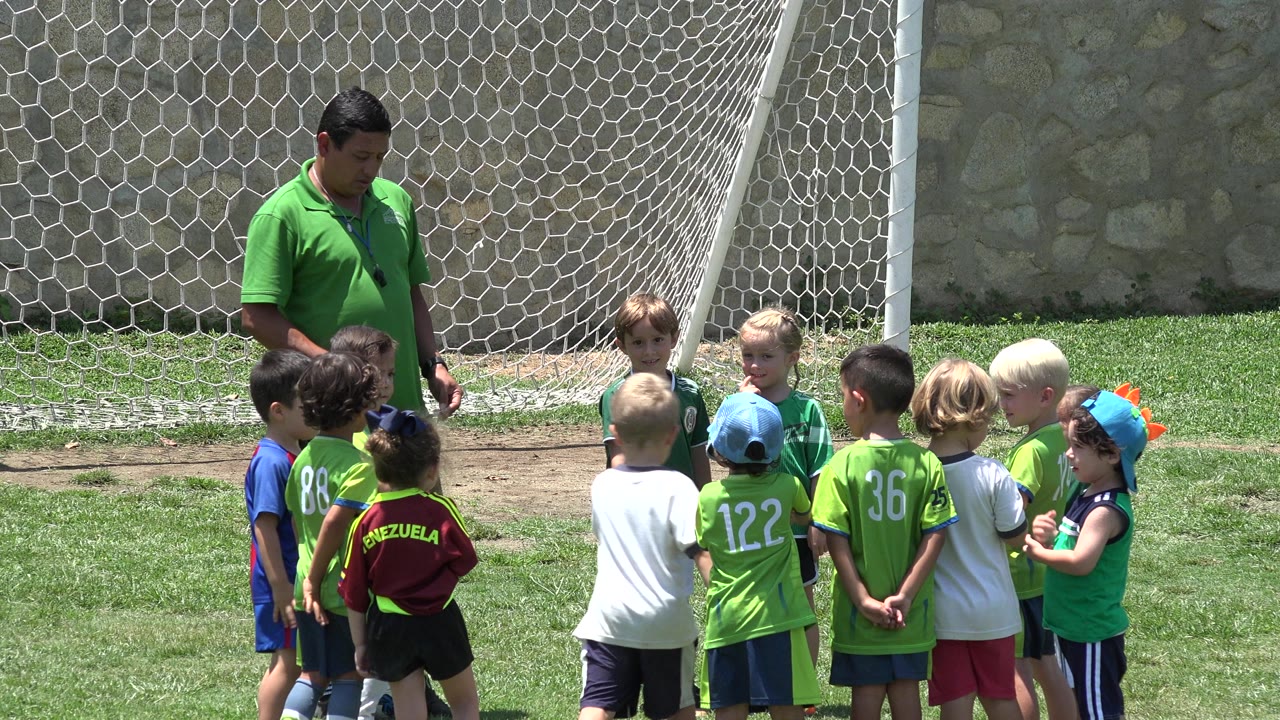 In Cabo San Lucas, Mexico, the group remains clustered on the grass while several children in bright green jerseys face the coach with the goal net behind them.