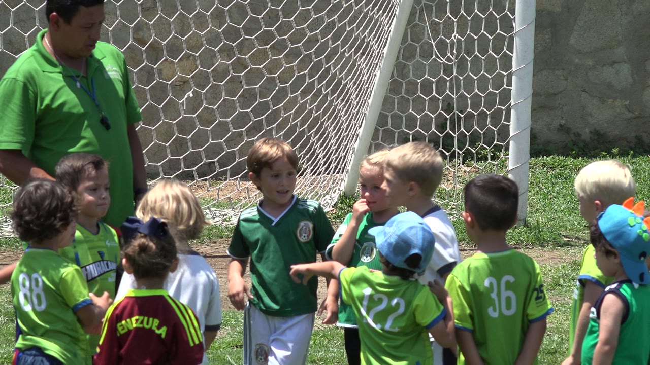 In Cabo San Lucas, Mexico, young children in green and white jerseys cluster around an adult coach beside a white goal net at the start of the clip.