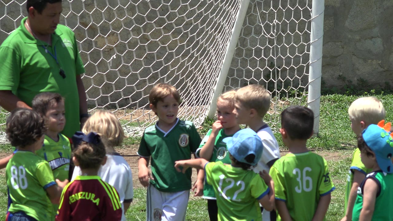 In Cabo San Lucas, Mexico, children in bright green jerseys stand with their backs and profiles to camera while a coach supervises near the goal net.