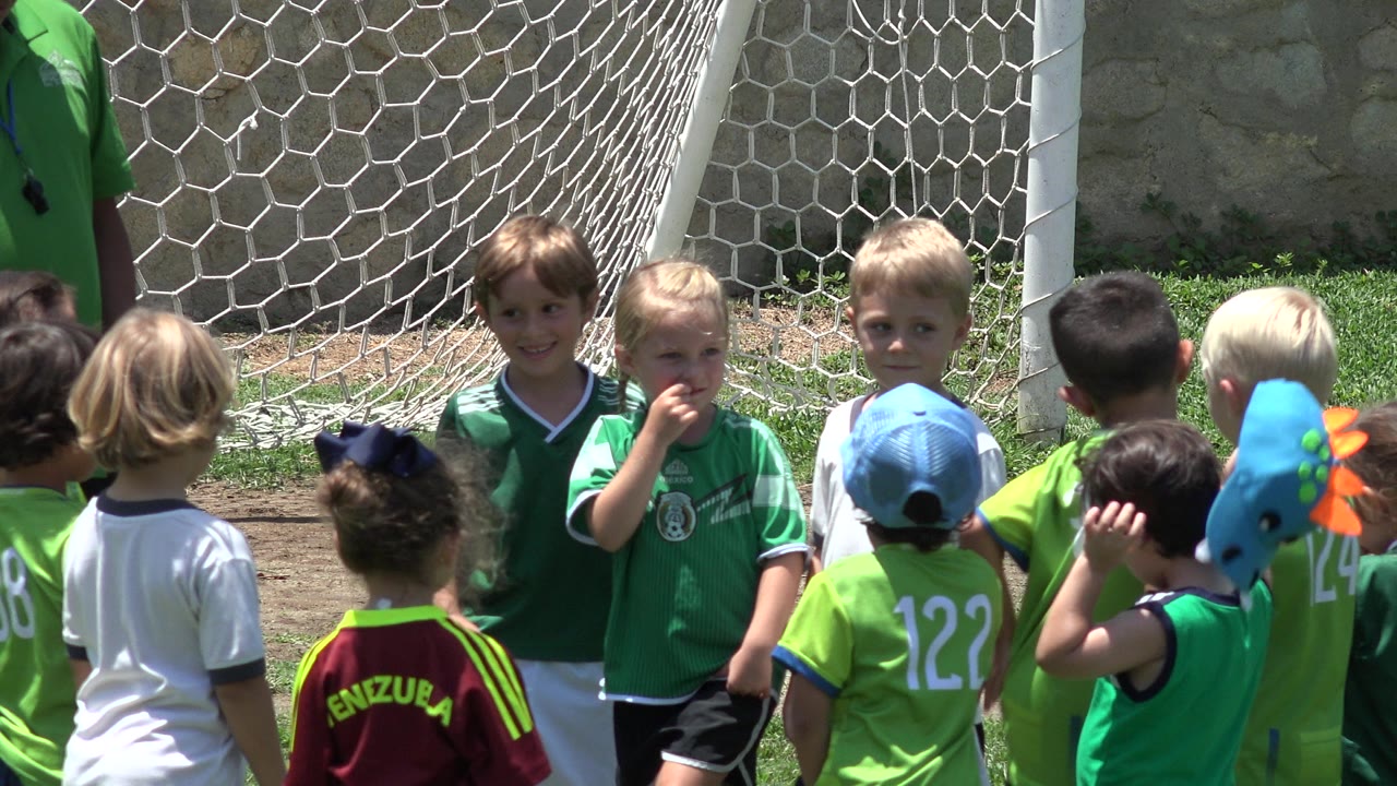 Several young children stand close together by the goal net, including a child in a light blue cap and others in green jerseys in Cabo San Lucas, Mexico.