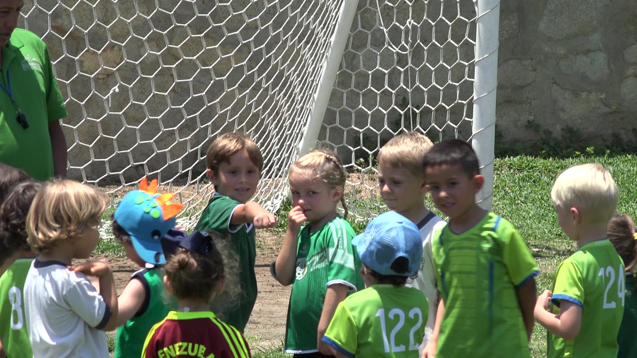 A group of young players in green and white shirts waits by the goal net in Cabo San Lucas, with one child pointing toward the camera side.