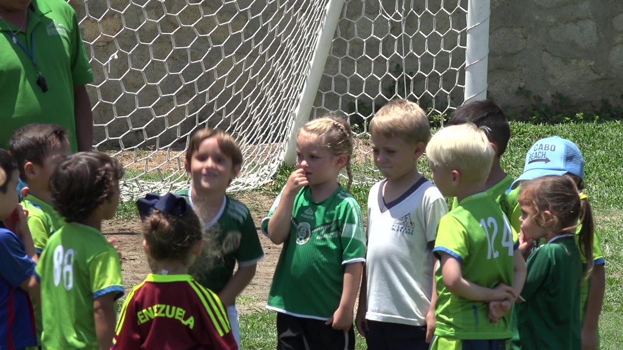 At 0:00, young children in green and white soccer jerseys stand close together on a grass field beside a white goal net in Cabo San Lucas, Mexico.