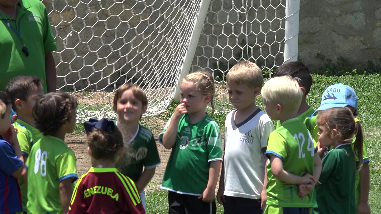 At 0:01, several children face different directions in bright sunlight near the goal net, with a blond child in a white jersey centered in the Cabo San Lucas soccer field scene.