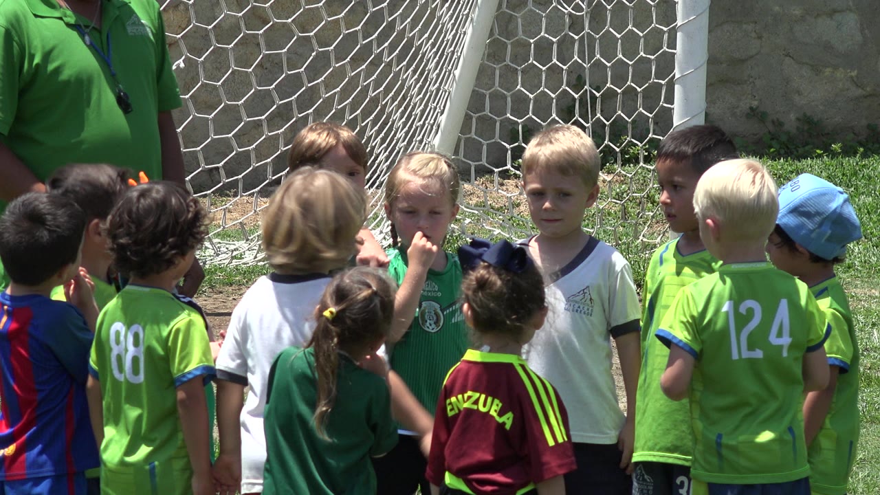 Young children in green and colorful soccer shirts stand near a white goal net in Cabo San Lucas, Mexico, with a partial adult visible at the edge of the group.