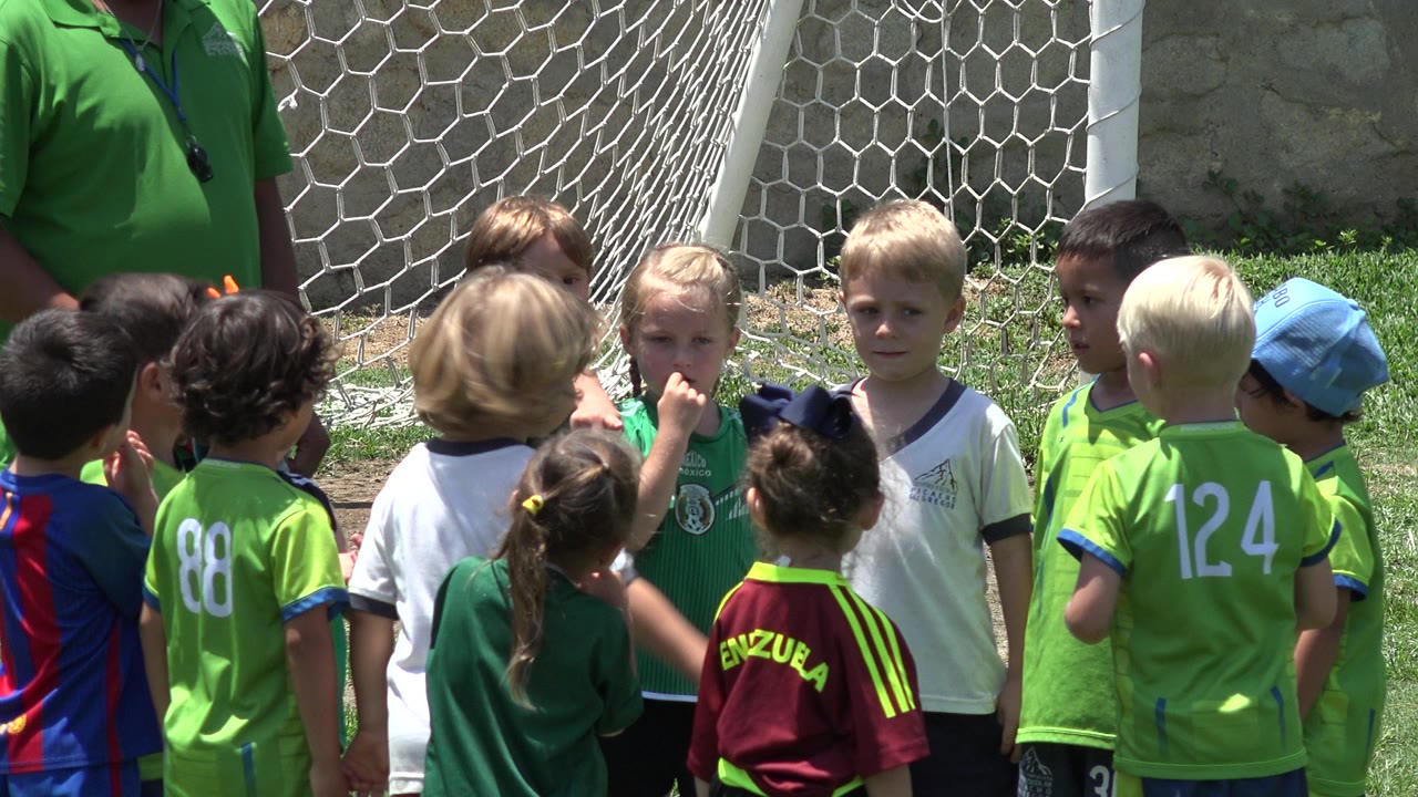 Children in green jerseys and casual sports shirts stand in a loose circle by the white goal net in Cabo San Lucas, Mexico, with several faces turned toward the center.